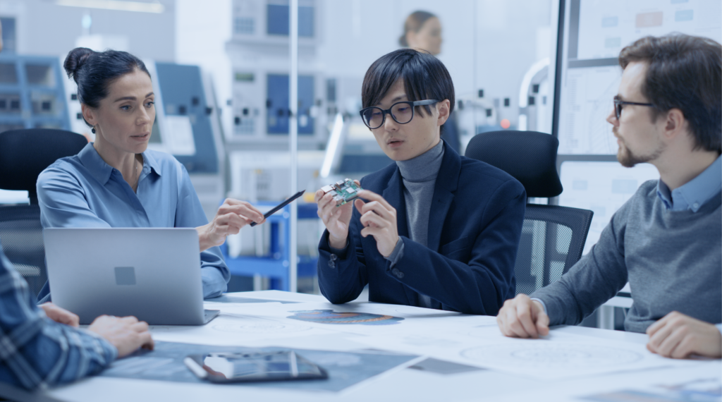 Diverse team in tech lab discussing circuit board chip over conference table.
