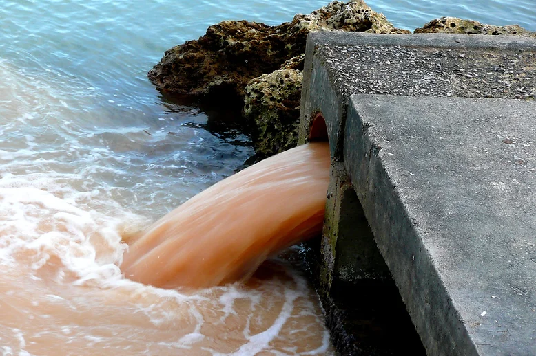 Polluted orange water pouring from drain pipe into ocean near rocks.
